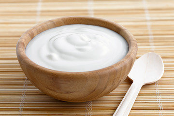 Wooden bowl of white yoghurt on bamboo matt with wooden spoon.