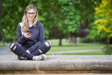 Obraz premium Young woman sitting on the wall in the park, writing a message on her mobile phone, park in the background