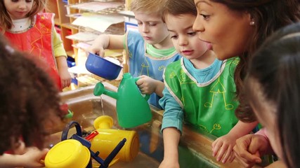 Nursery children playing at the water table in their classroom. The teacher is talking to them. - Powered by Adobe