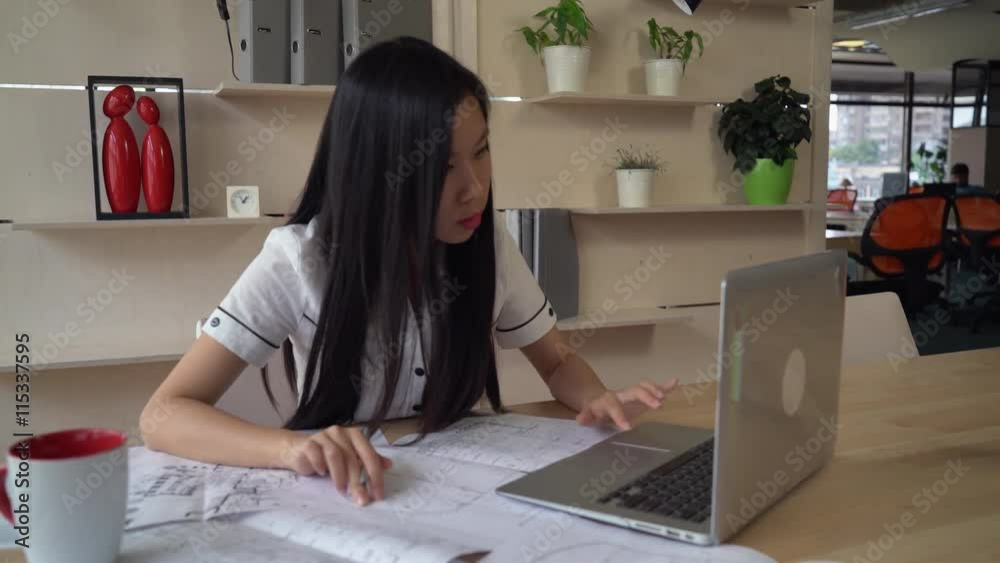 Poster Female looking on blueprint and checking with electronic version on computer. Asian young professional designer works at the working place with table laptop . Multicultural smiling worker wearing in