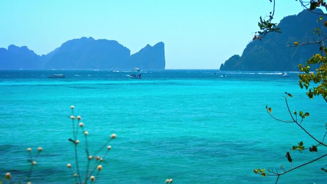 Heavy boat traffic criss-crosses the bay near a popular tourist beach in Thailand, with mountains and seacliffs in the background.