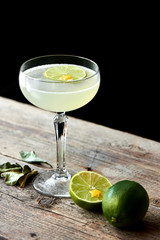 The glass of alcoholic cocktails Daiquiri consists of rum on the leaves of lime, elderflower syrup stands on wooden table on black background