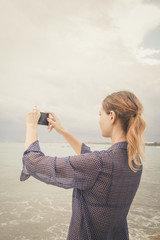 Woman taking photos of the beach with cell phone during the cloudy day