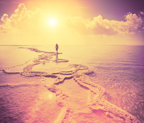 Fototapeta na wymiar Silhouette of young woman walking on Dead Sea salt shore at sunrise towards the sun