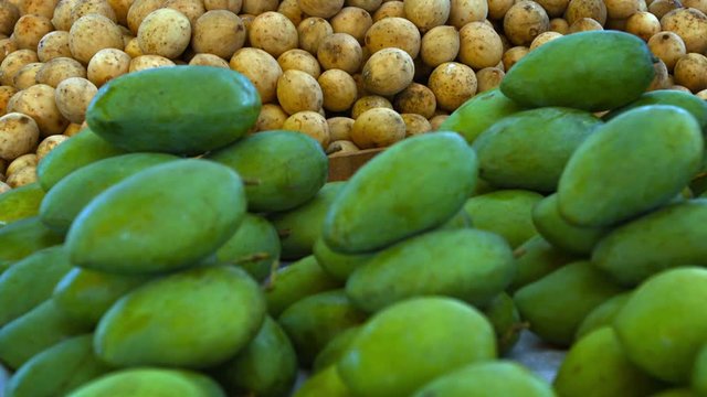 Video 1080p - Small, green mangoes, harvested early for their sour flavor, and sweet lanzones fruit, piled high for sale at an outdoor, public market in Asia.
