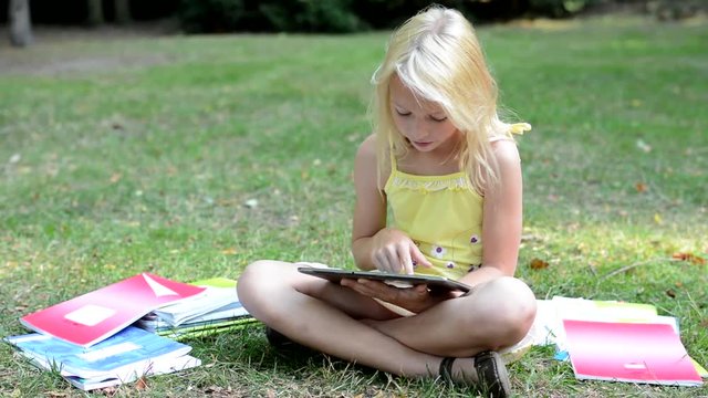 little smart girl sits in the park and searchs something on the tablet - she thinks and writes - surrounded by workbooks