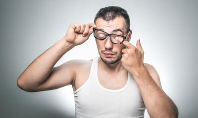 Man wiping eyes with his fingers, isolated on gray background. Waking morning. Guy with undershirt holding eyeglasses