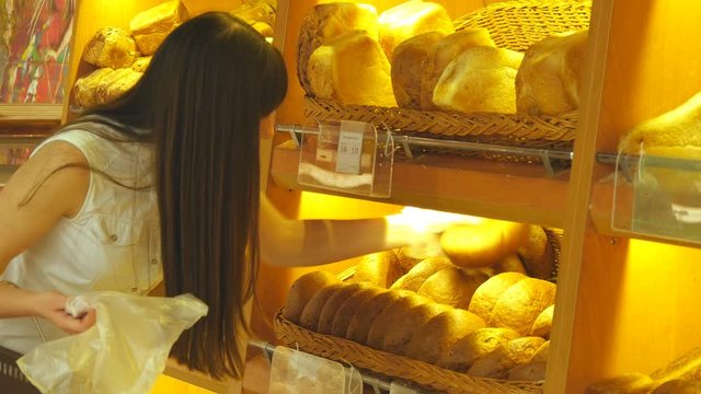 Woman chooses and putting a fresh bread loaf into a package in the supermarket. Young girl taking a loaf of bread from the shelf, smelling it and putting it into the basket. Shopping in the grocery