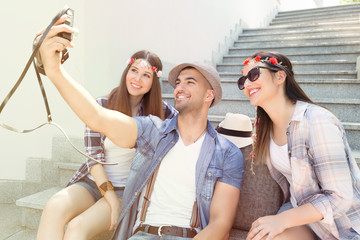 Three young friends are enjoying their summer vacation. They are sitting on the outdoor steps while young man is taking a self-portrait with two girls.