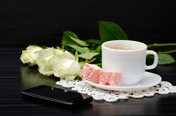 Side view of a coffee cup with milk, oriental sweets. smartphone, white roses on a black background
