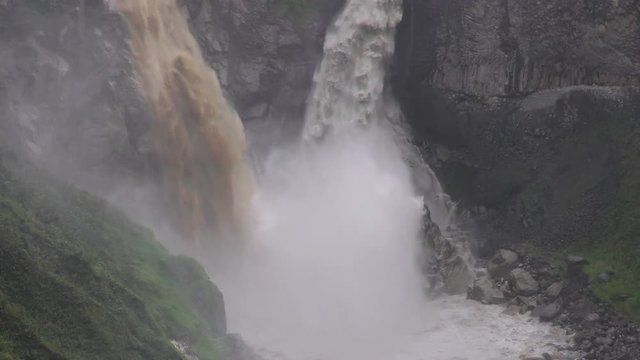 A dynamic view of a powerful waterfall plunging down a rugged cliff face near Banos de Agua Santa, Ecuador, dense mist at its base and dark moss-covered rock around the untamed scene.