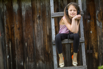 Little naughty girl with headphones sitting on a wooden stepladder in the village outdoors.