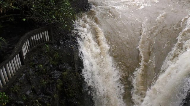 A high-angle view of turbulent muddy water rushing over dark wet rocks at the Pailon del Diablo near Banos, Ecuador, white foam and rapids, a stone railing and lush green foliage on the left bank.