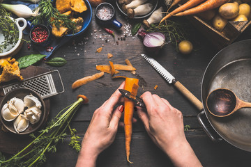 Female woman hands peeling carrots on dark wooden kitchen table with vegetables cooking ingredients, spoon and tools, top view