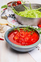 Bowl with  tomatoes sauce on kitchen table with cooking pot and green vegetables, close up