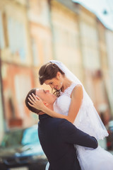 Fototapeta premium Wedding couple walking cheerfully in urban landscape. Bride and groom embracing and kissing. People in love. City streets. Wedding day. Horizontal color photo.