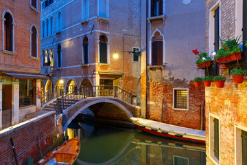 Fototapeta premium Lateral canal and pedestrian bridge in Venice at night with street light illuminating bridge and houses, with docked boats, Italy
