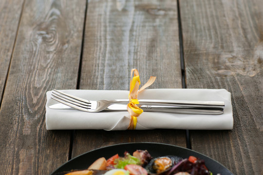 Cutlery on brown wooden background with seafood, void. Horizontal position of white napkin with knife and fork, seafood meal in downside of image, copyspace on wood. Focus on restaurant cutlery