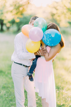 Young happy family and little son hide theis faces by colorful baloons. Green meadow on background.
