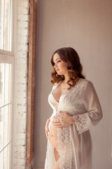 Naklejka premium Beautiful pregnant woman in white peignoir looks out the window and gently hugs her belly in the light interior.