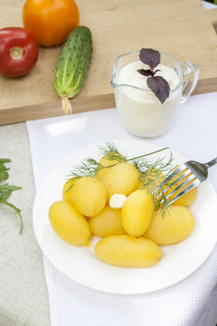 Boiled potatoes with dill and butter in white plate. Fresh cucumber and tomatoes on wooden board. Sour cream in sauce boat with basil. 