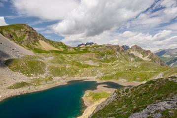 Fototapeta na wymiar High altitude blue lake in idyllic uncontaminated environment once covered by glaciers. Summer adventures and exploration on the Italian French Alps. Expansive view from above, dramatic sky.