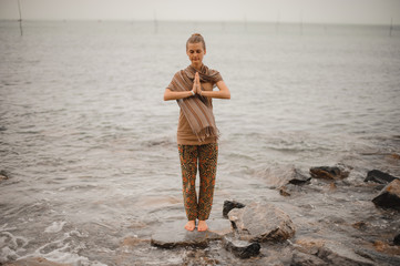 Woman standing a grateful namaste yoga pose on the beach next to the ocean in cloudy weather. Zen, meditation, peace.