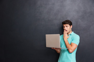 Amazed shocked young man standing and using laptop