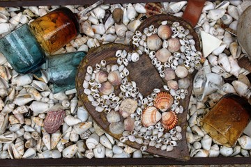 Composition of heart made of wood and shells surrounded by broken colorful glasses. At the background many shells. - obrazy, fototapety, plakaty