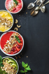 Healthy summer breakfast concept. Colorful fruit smoothie bowls with nuts, oat granola and mint leaves, five silver spoons on black background. Yellow, pink, red, green. Top view, copy space