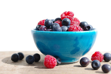Juicy mature berries of raspberry and bilberry in a small blue bowl on a wooden surface.