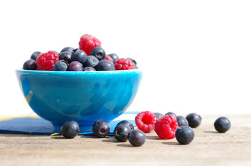 Juicy mature berries of raspberry and bilberry in a small blue bowl on a wooden surface.
