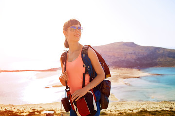 female traveler with backpack and a solar panel standing on the trail against sea and blue sky at early morning