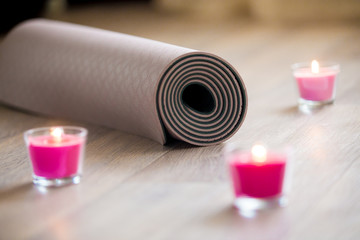 Rose-colored lighted pink wax candle and rolled brown yoga, pilates mat on the floor ready for...