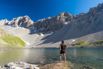 Fototapeta na wymiar Hiker relaxing at high altitude blue lake in idyllic uncontaminated environment once covered by glaciers. Summer adventures and exploration on the Italian French Alps. Clear blue sky.