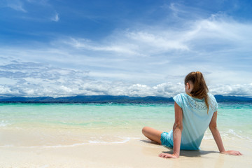 Fototapeta na wymiar young beautifull girl is sitting on the white sand of tropical beach near turquoise sea under cloudy sky