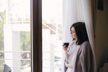 Young beautiful woman in a sweater drinking red wine near big window in a cozy room