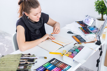 Young female artist drawing sketch using sketchbook with pencil at her workplace in studio. Side view portrait of inspired painter.