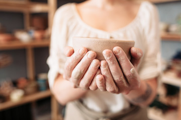 Woman messy hands holding bowl, closeup, blurred background, focus on potters palms with pottery