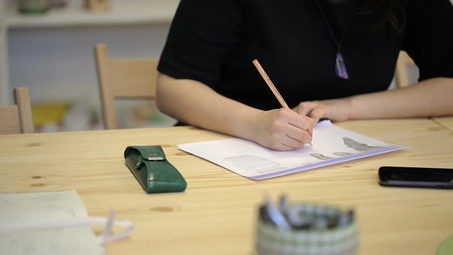 On wooden table is cellphone, eyeglass case, paper sheet on which woman draws.