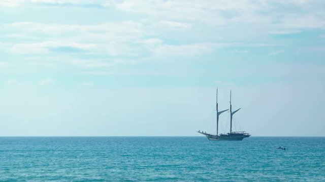 Two masted, wooden sailing yacht, anchored on the horizon off a popular tropical beach in Southeast Asia.