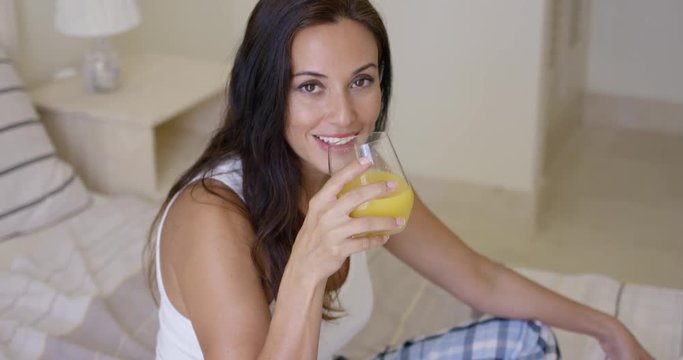 Smiling healthy young woman drinking a glass of freshly squeezed orange juice as she sits on her bed in the morning