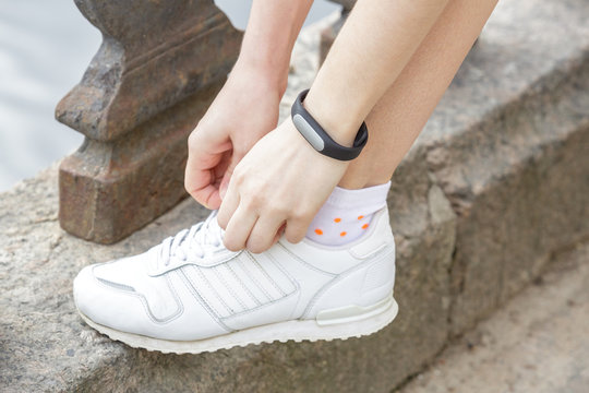 Woman hands with black fitness bracelet adjusts laces on white sneakers. On the street, on the parapet of the river.
