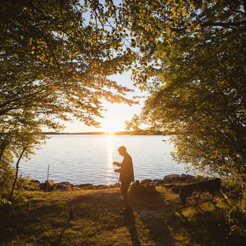 Sweden, Skane, Immeln, Mid adult man standing with fishing rod by river at sunset