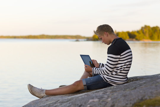 Sweden, Ostergotland, Teenage boy (16-17) sitting on rock and using digital tablet