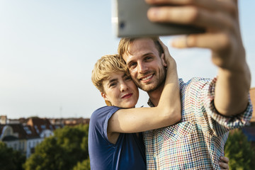 Germany, Berlin, Young couple hugging and taking selfie with smart phone