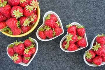 Fresh picked strawberries in a yellow bowl, and white heart shaped bowls, on a gray background
