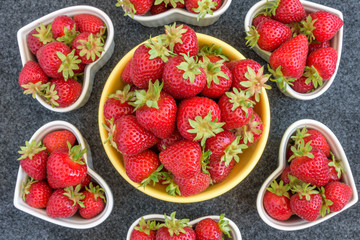 Fresh picked strawberries in a yellow bowl, and white heart shaped bowls, on a gray background
