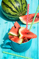 Fresh watermelon slices in a cup with colored straws, blue wooden background