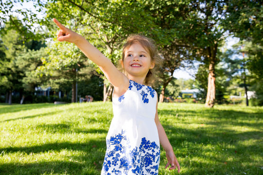Beautiful kid having fun in green summer park, laughing. Warm sunny weather, healthy child, happiness, freedom, emotions. Cute girl reaching finger, pointing.